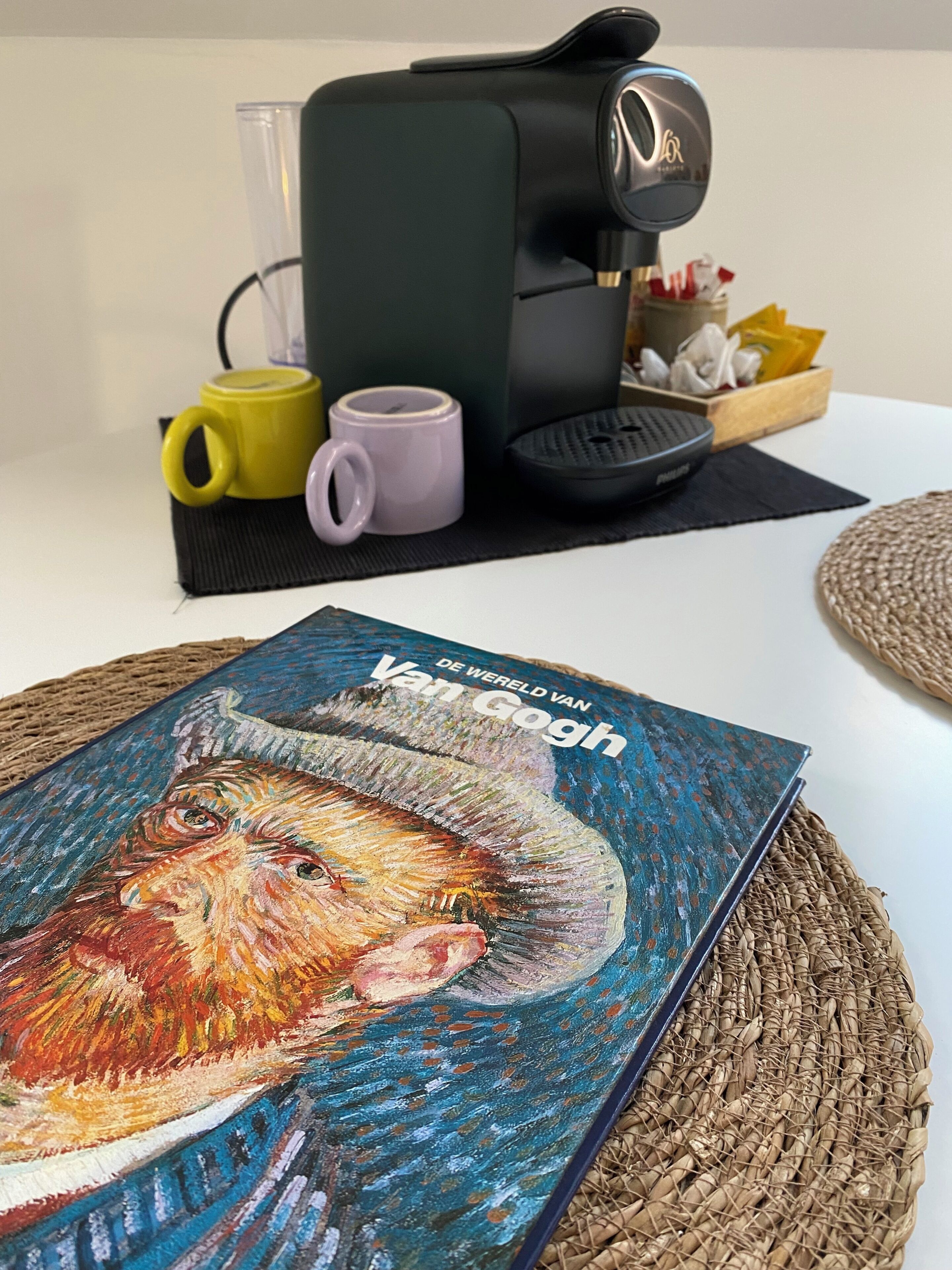 A coffee maker, two mugs, a woven placemat, and a book titled 'Van Gogh' on a table.