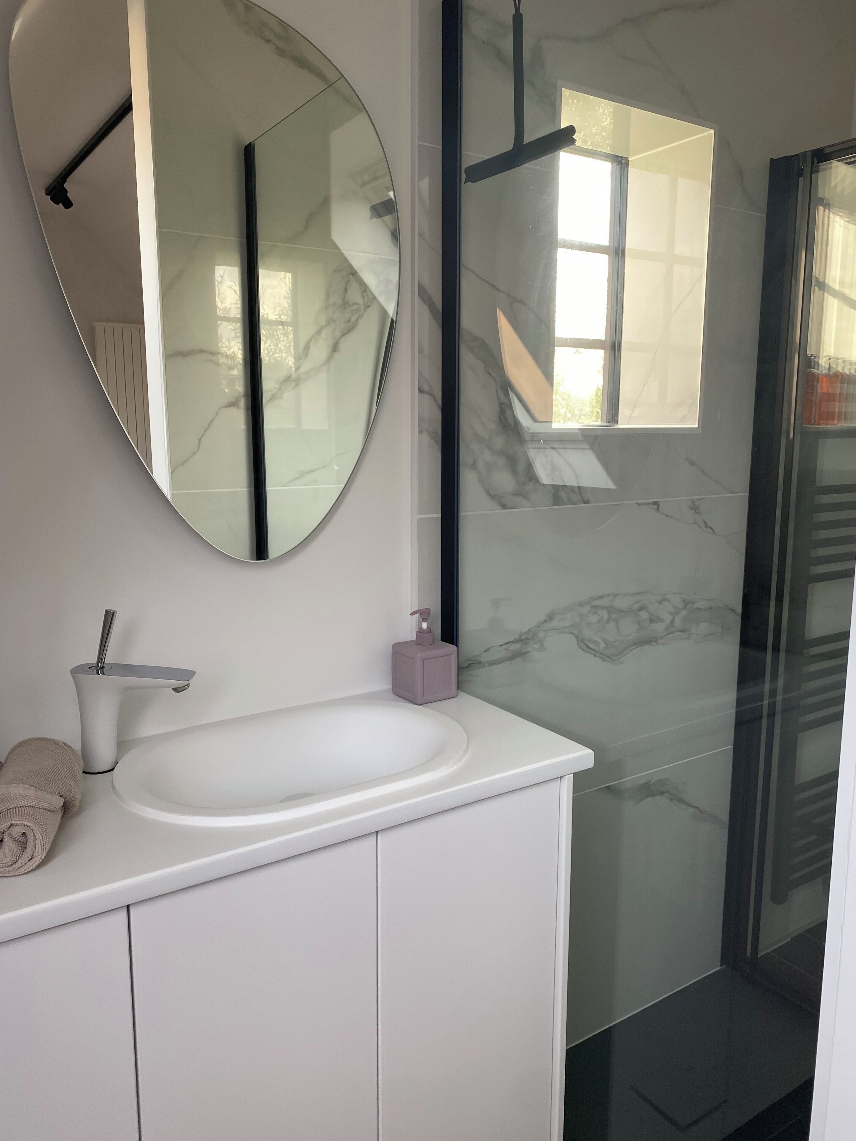 Een badkamer met een wit wastafelmeubel, een ronde spiegel, een witte wastafel, een handdoek en een glazen douchecabine.
