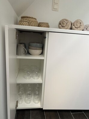 A white cabinet with shelves containing bowls, a kettle, and wine glasses.