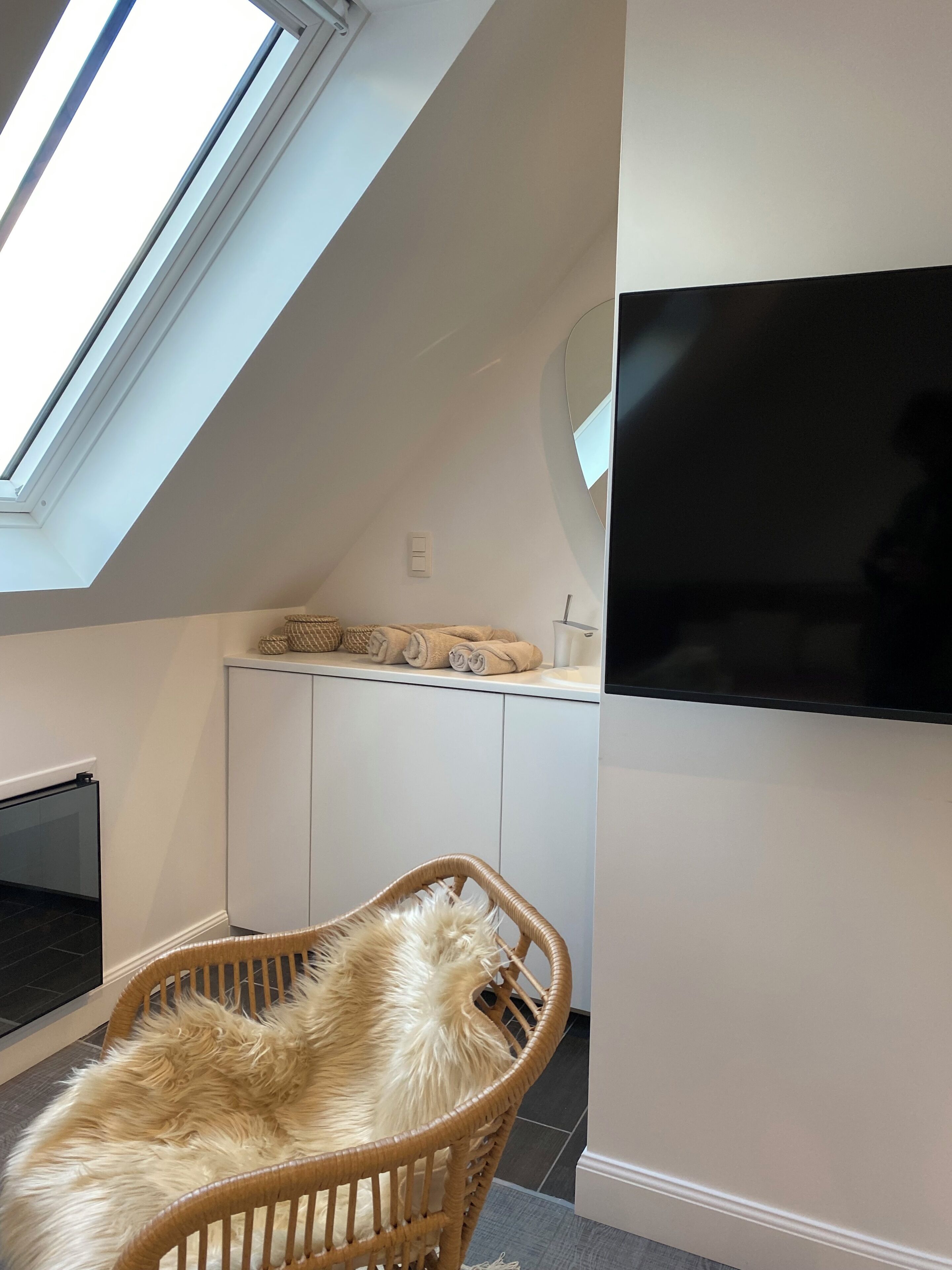 A room with a high ceiling, a skylight, a flat-screen TV mounted on the wall, and a wicker chair with a fluffy cushion.