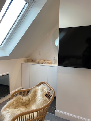 Una habitación con techo alto, tragaluz, televisor de pantalla plana en la pared y una silla de mimbre con cojín mullido.