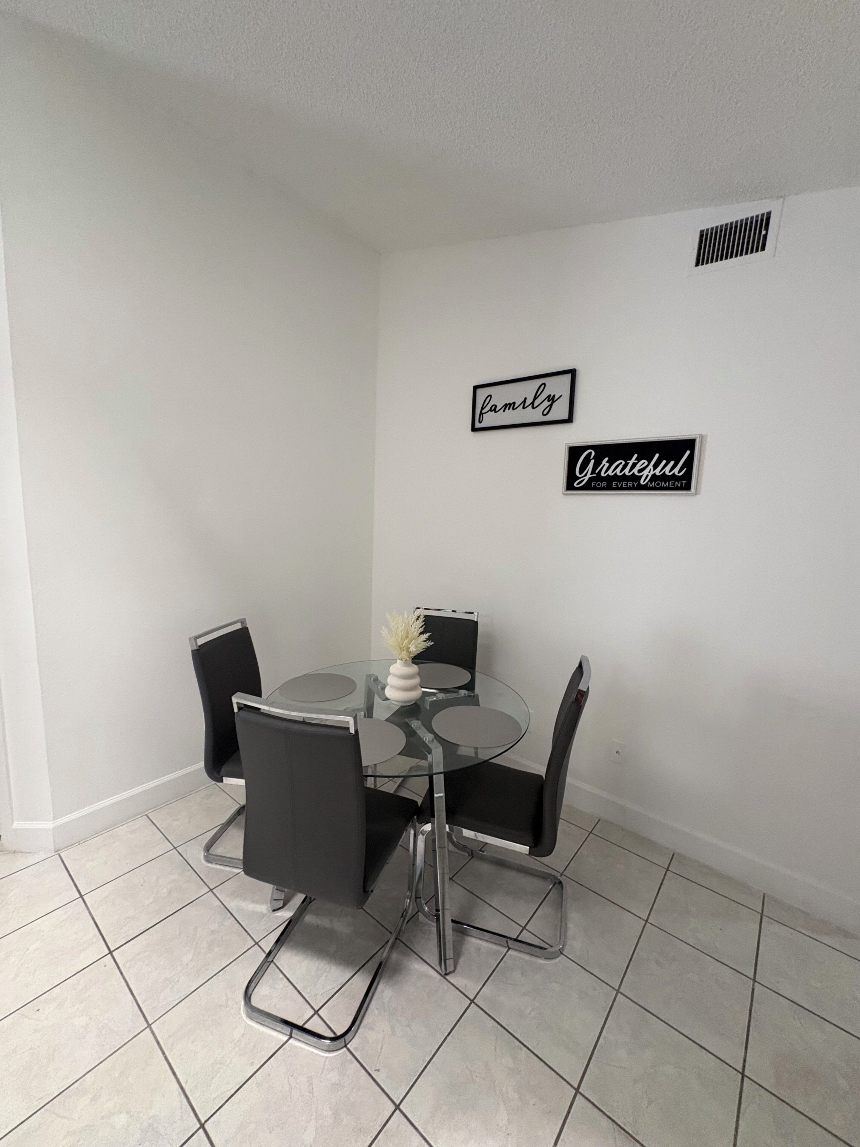 A dining area with a glass table and four chairs, two wall decorations, and a tiled floor.