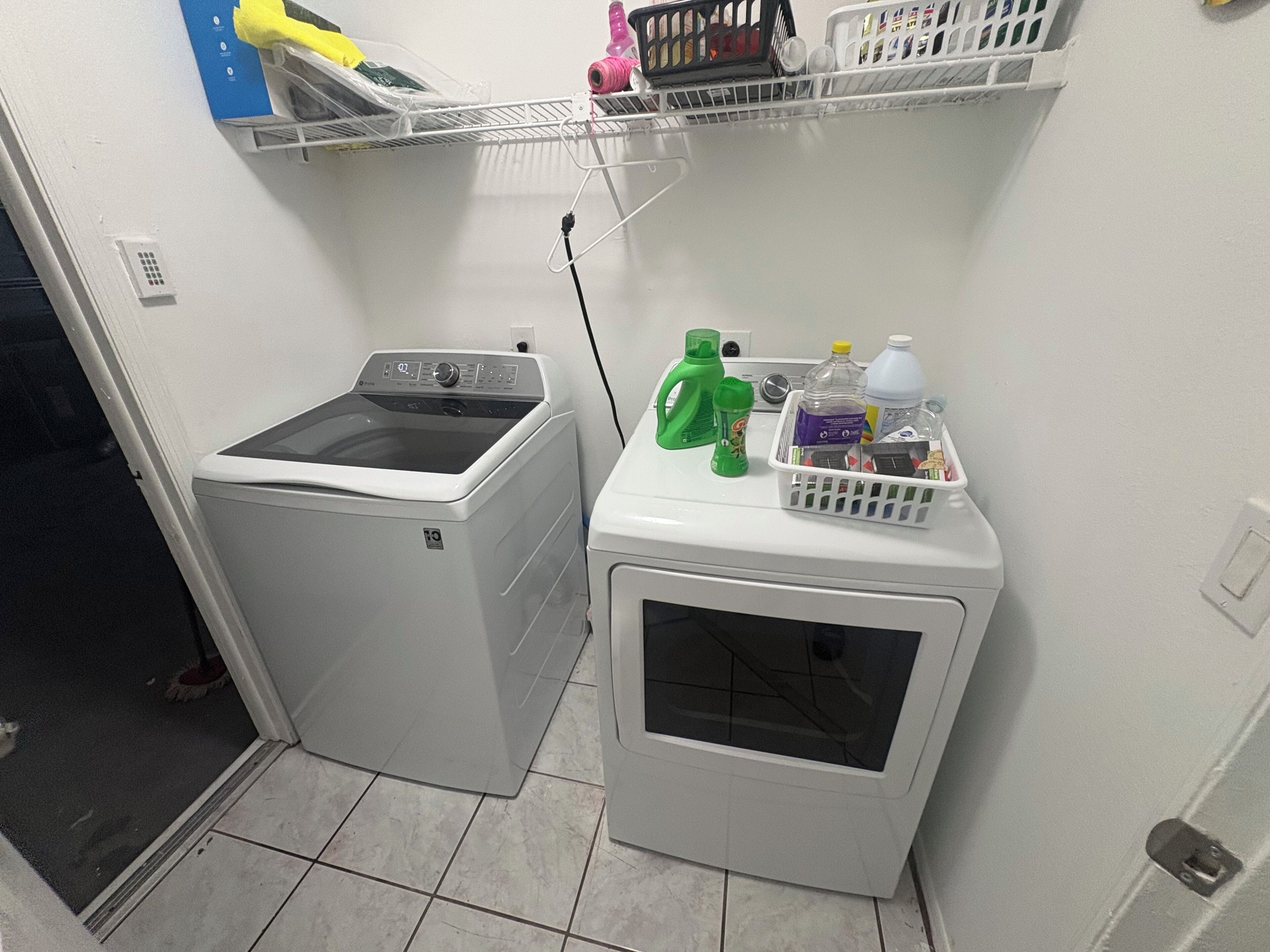 A laundry area with a washing machine and dryer, a shelf with laundry supplies, and a door leading to another room.
