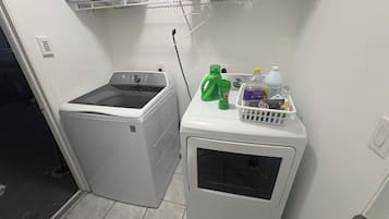 A laundry area with a washing machine and dryer, a shelf with laundry supplies, and a door leading to another room.