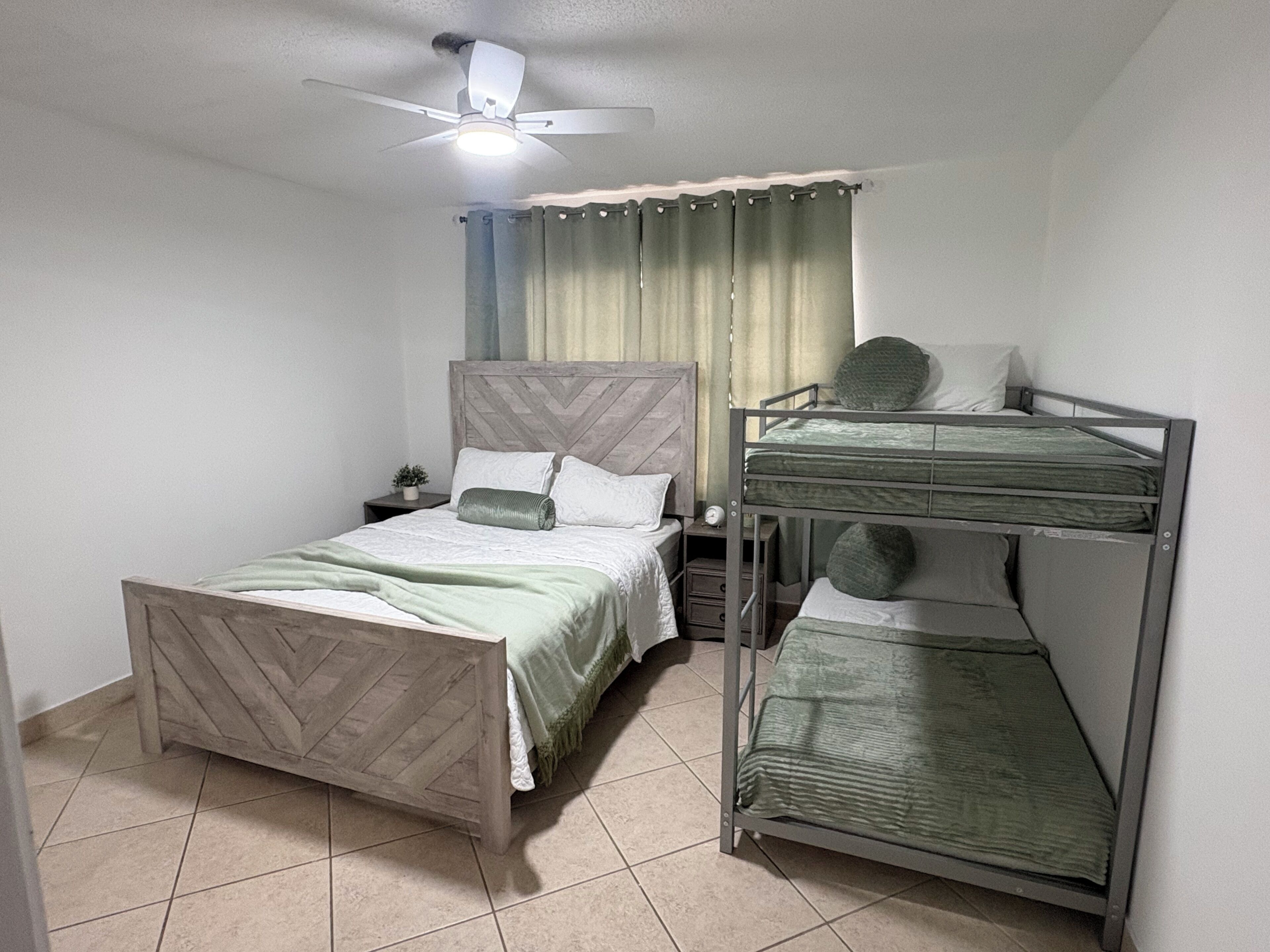 A room with a bed, a bunk bed, a ceiling fan, a nightstand, a potted plant, and a window with curtains.