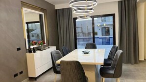 Ein stilvolles Esszimmer mit einem rechteckigen Tisch, sechs Stühlen, einer Pendelleuchte, Blick auf einen Swimmingpool sowie einer Küchenzeile mit einem weißen Schrank und einem Fenster.