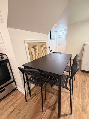 Un comedor con una mesa y cuatro sillas, una ventana con persianas, una puerta y una zona de cocina con horno.