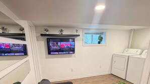 A living room with a grey sofa, a wooden coffee table with a plant, a TV mounted on the wall, and a laundry area with washing machines. - Room to Stay in Niagara Falls (Niagara Falls)