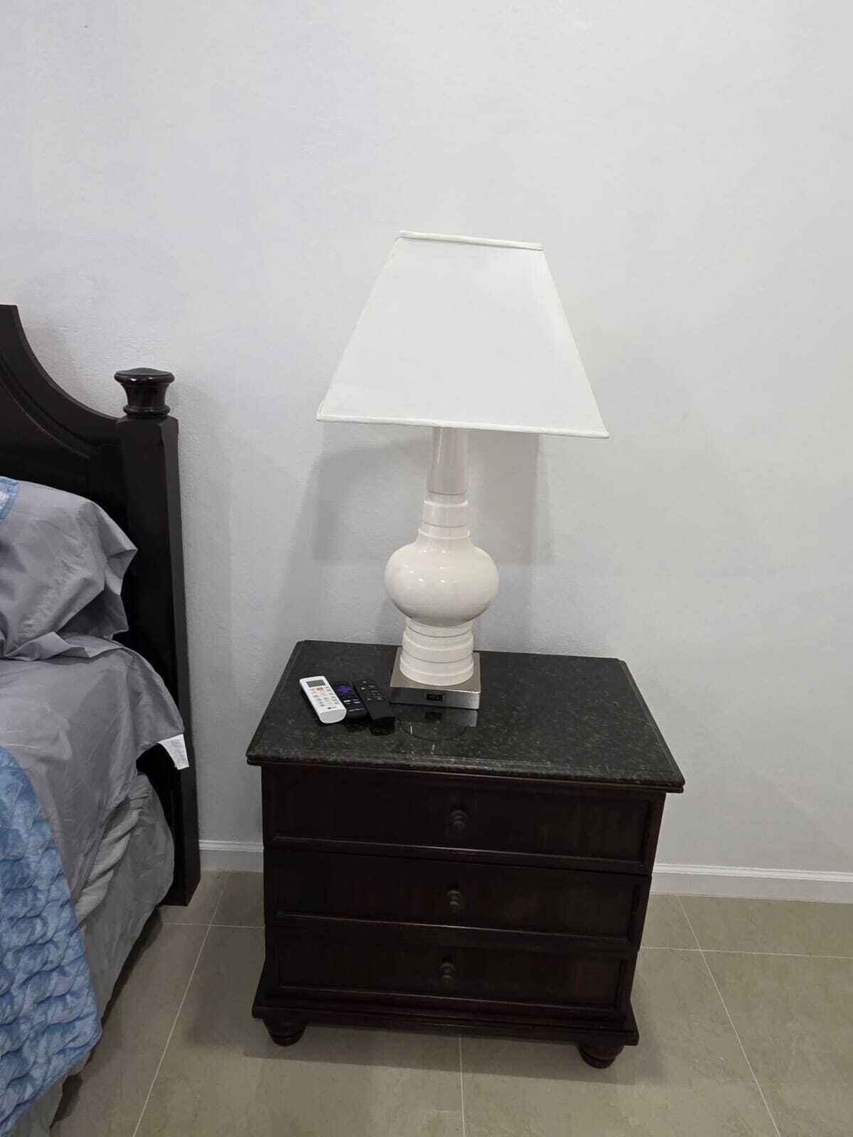 Een donker houten nachtkastje met een witte lamp, een afstandsbediening en een klein elektronisch apparaat.