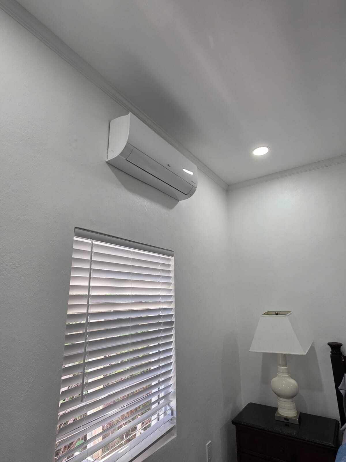 Une chambre avec une fenêtre équipée de stores, un climatiseur fixé au mur, une table de chevet avec une lampe et un luminaire au plafond.