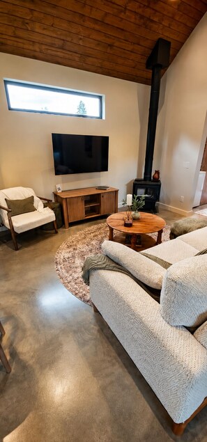 A modern living room with a large sofa, a flat-screen TV, a wooden coffee table, and a black wood-burning stove. - Boutique Retreat • Private Sauna • 2 King Suites (West Kelowna)