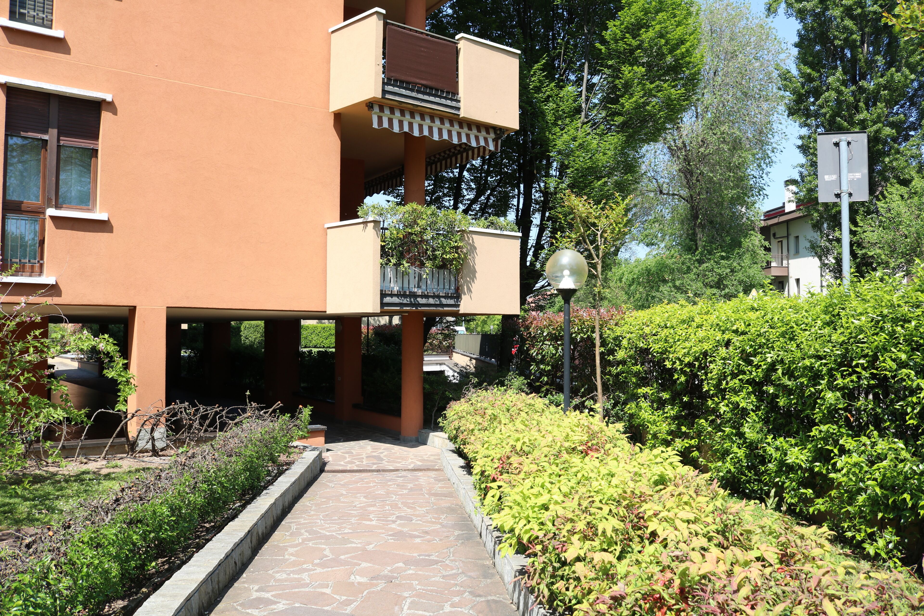A pathway leading to a building with a balcony, surrounded by greenery and a street lamp.