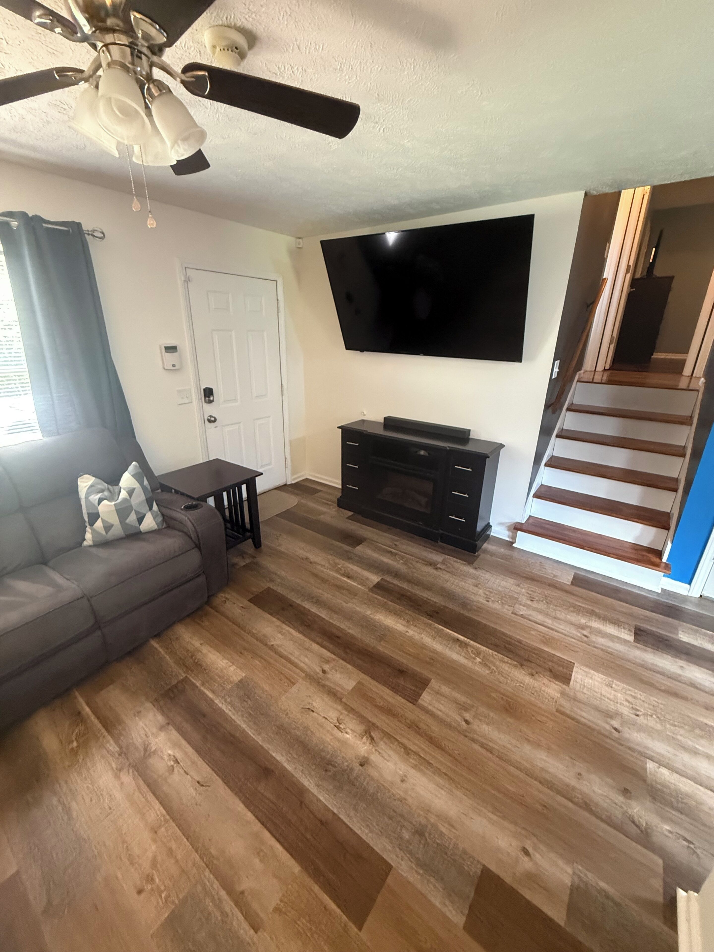 Un soggiorno con ventola a soffitto, televisore a schermo piatto montato sulla parete, mobile TV in legno scuro, divano grigio con cuscino geometrico e una scala che conduce a un piano superiore.