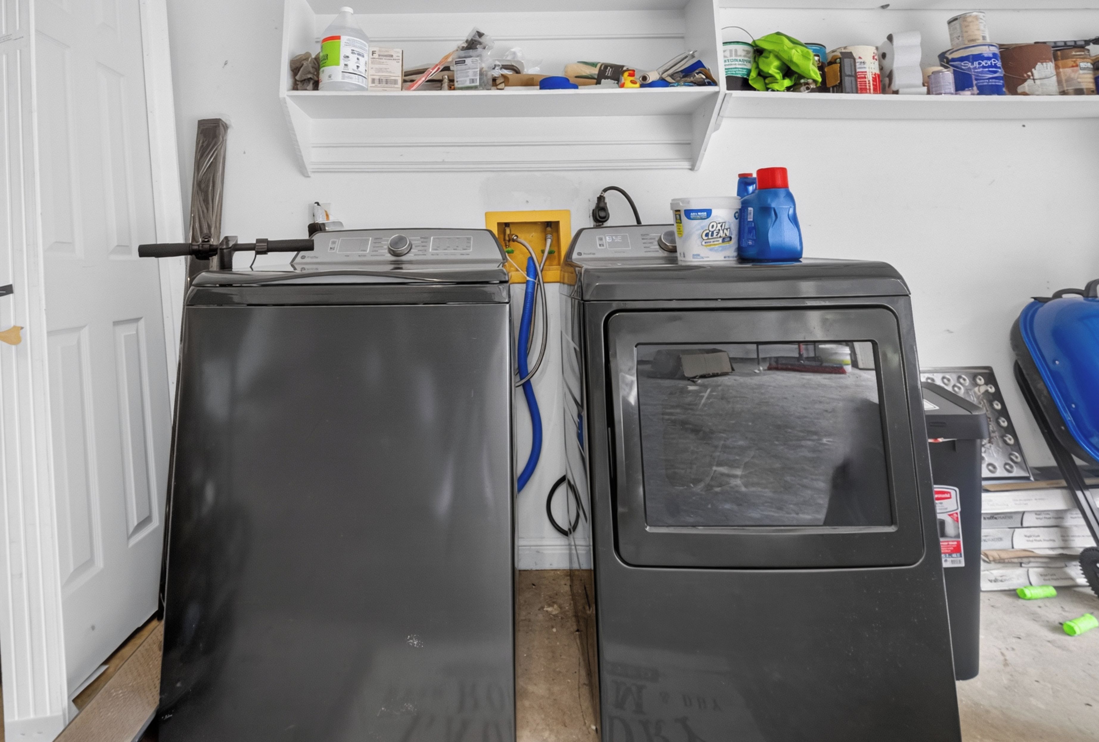 Dos lavadoras una encima de otra en una habitación con una puerta blanca y un estante superior con varios objetos.