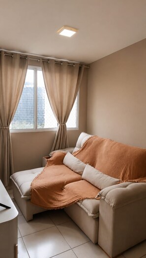 A room with a beige sofa, a white bedside table, and a window with curtains. - Cozy Apartment in Barra Olímpica | 10 min from Recreio + Rio Centro (Rio de Janeiro)