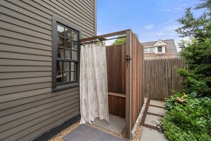 Eine Umzäunung für eine Außendusche aus Holz mit Fenster, eine Holzzahlung und ein Gartenbereich mit trittfesten Steinen.
