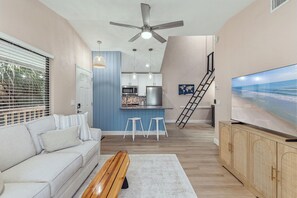 A modern living room with a sofa, a wooden bench, a TV, and a kitchen area with a blue accent wall. - 🌊 Beachy Dreams | Non-Drive Beach NSB | Arcade + Paddleboard + Gear (New Smyrna Beach)