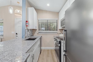 A modern kitchen with granite countertops, stainless steel appliances, and a window with blinds. - 🌊 Beachy Dreams | Non-Drive Beach NSB | Arcade + Paddleboard + Gear (New Smyrna Beach)