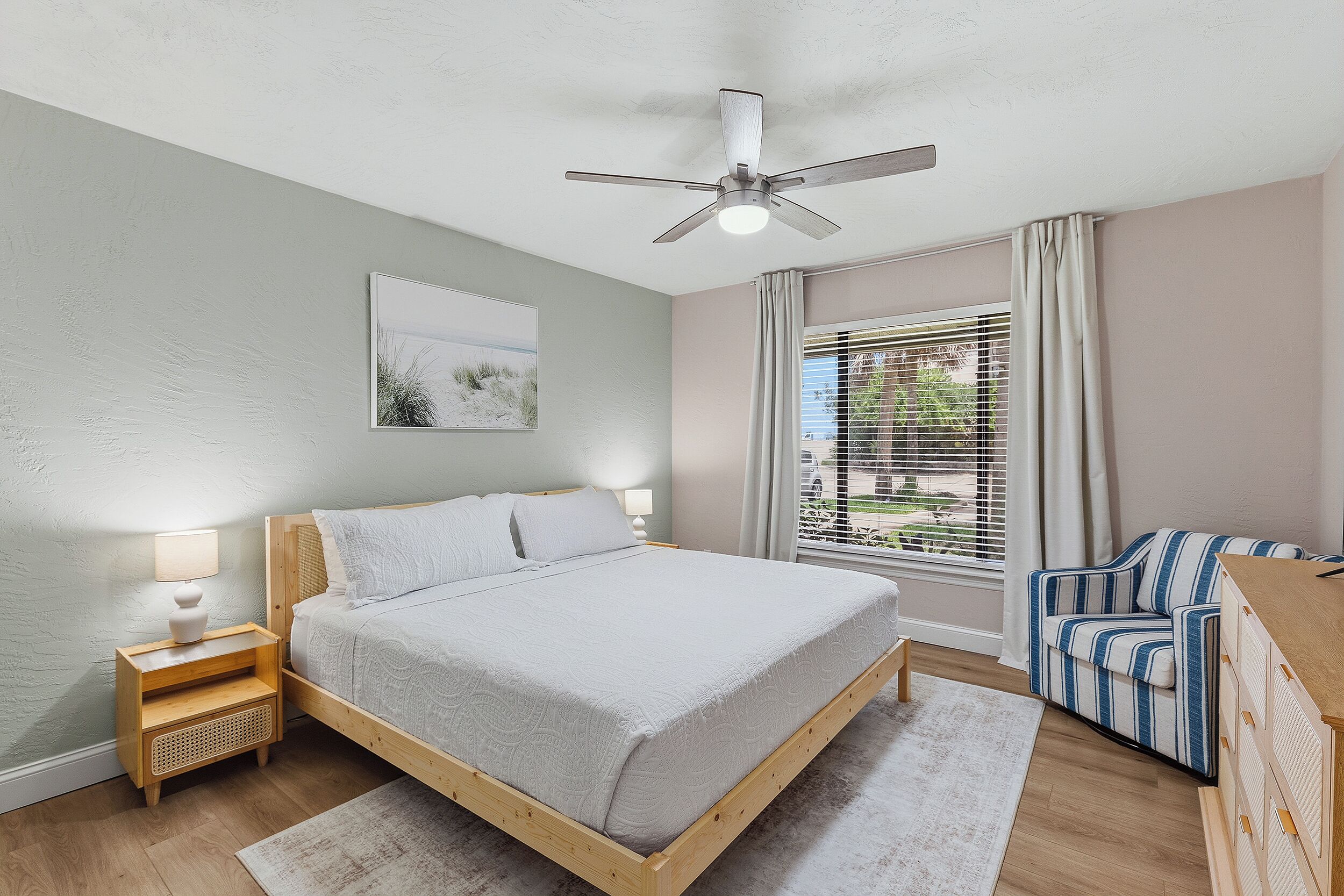 A bedroom with a bed, bedside tables, a ceiling fan, a window with curtains, and a view of the outdoors.