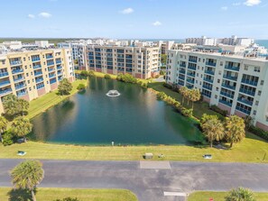 A residential area with a central pond, surrounded by multi-story buildings with balconies. - 🌊 Oceanwalk 15-503 | Non-Drive Beach NSB | Soaking Tub + 2 Pools (New Smyrna Beach)