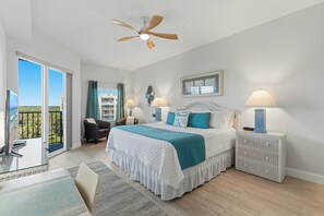 A bedroom with a bed, two bedside lamps, a ceiling fan, a view of the outdoors, and a balcony. - 🌊 Oceanwalk 15-503 | Non-Drive Beach NSB | Soaking Tub + 2 Pools (New Smyrna Beach)