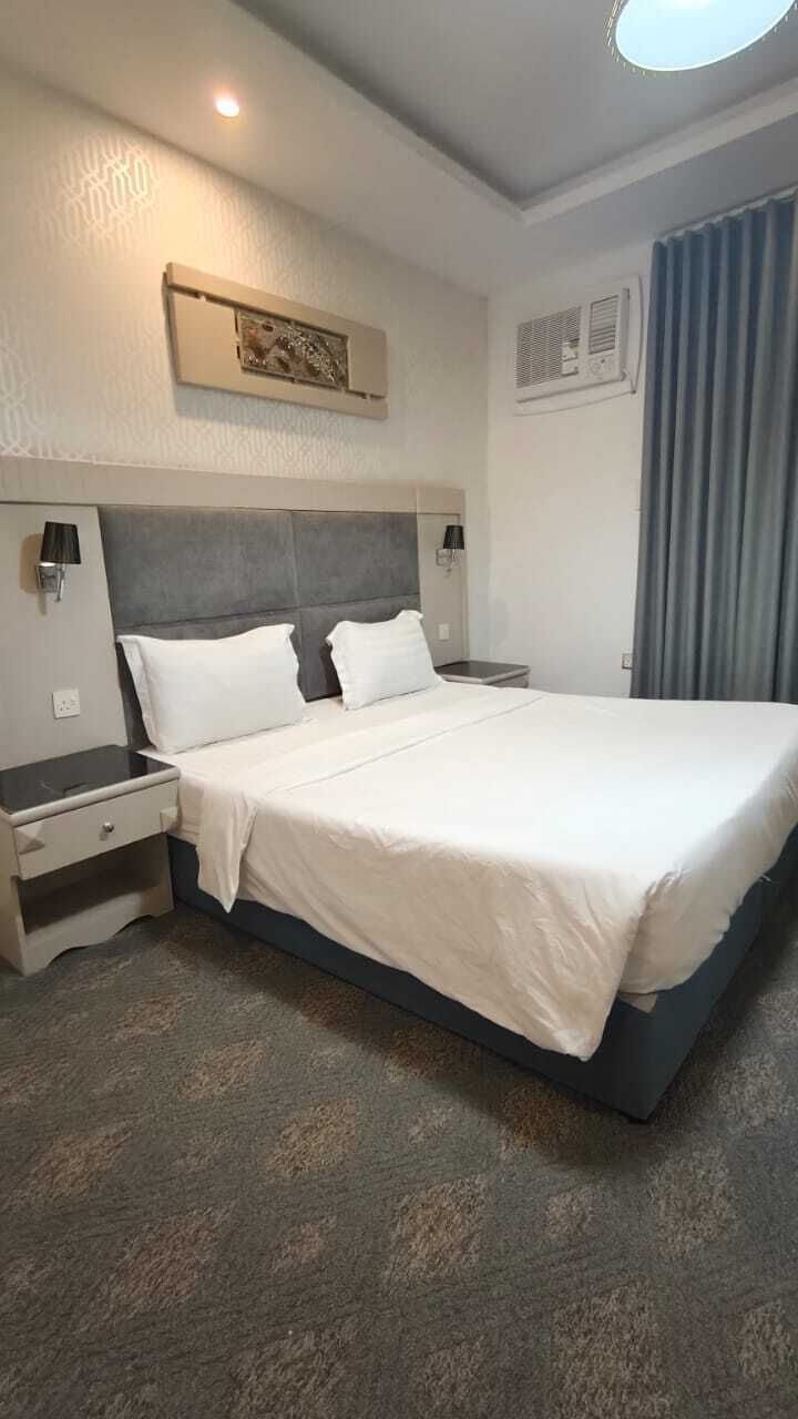 A hotel room with a large bed, bedside tables, a headboard, wall-mounted lamps, a ceiling light, and a window with curtains.