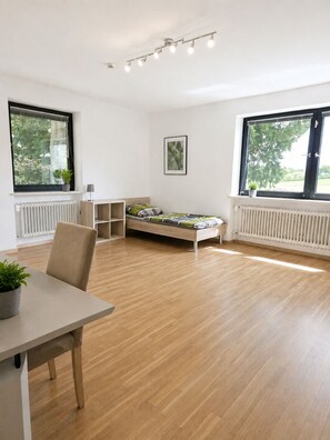 A modern room with a wooden floor, a bed with a green and white bedspread, a small table with a chair, and two windows with blinds. - Greengarden Apartment for Construction Workers (Vilsheim)