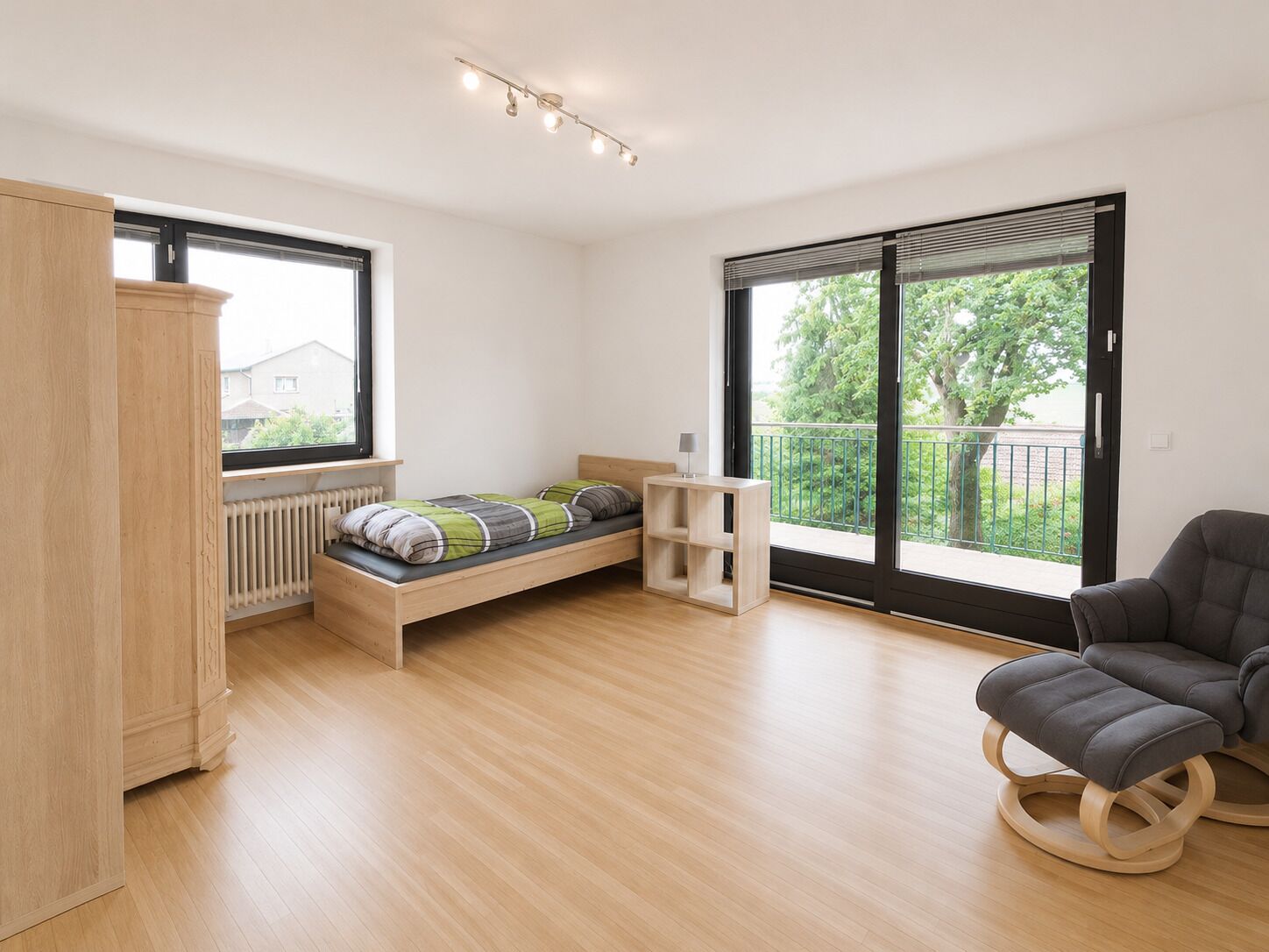 A modern bedroom with a wooden bed, a grey armchair, and a view of greenery through large windows.