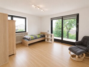A modern bedroom with a wooden bed, a grey armchair, and a view of greenery through large windows. - Greengarden Apartment for Construction Workers (Vilsheim)