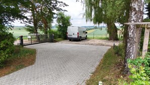 En asfalterad uppfart leder fram till en grindad entré, där en skåpbil står parkerad. Området är omgivet av träd och det finns utsikt över en grön äng.