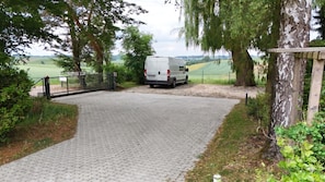 A paved driveway leading to a gated entrance with a parked van, surrounded by trees and a view of a green field. - Greengarden Apartment for Construction Workers (Vilsheim)