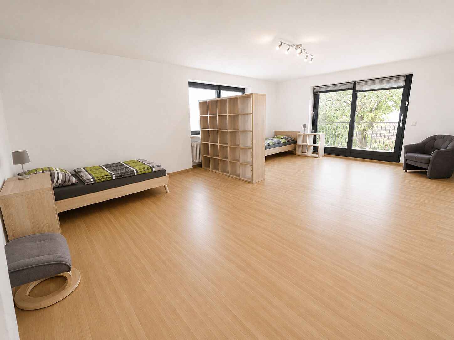 A modern, well-lit room with a wooden floor, a sofa, a bed, a bookshelf, and a chair.