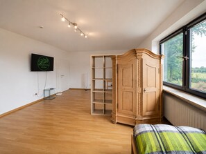 A room with a wooden floor, a large window with a view of greenery, a wooden cabinet, and a television mounted on the wall. - Greengarden Apartment for Construction Workers (Vilsheim)