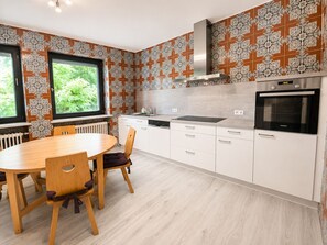 A modern kitchen with a dining table, chairs, a stove, and a window with a view of greenery. - Greengarden Apartment for Construction Workers (Vilsheim)
