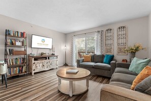Ein Wohnzimmer mit einer Couch, einem Couchtisch, einem Bücherregal, einem Fernseher und einem Fenster mit Vorhängen.