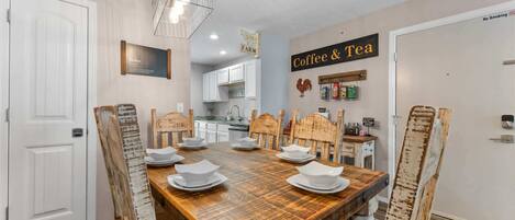 A dining area with a wooden table set for six, surrounded by rustic chairs. A kitchen is visible in the background, and a 'No Smoking' sign is on the wall.