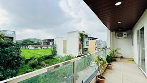 A balcony with a view of a green field and buildings, a glass railing, and potted plants.