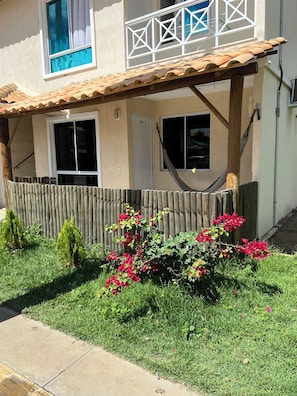 Un edificio de dos pisos con cerca de madera, una hamaca y un jardín con flores rojas.