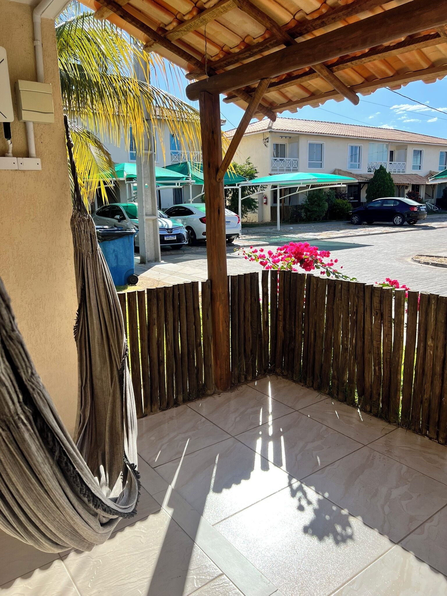 Un espacio al aire libre techado con piso de baldosas, cercado por una valla de madera, y una vista de coches estacionados y edificios al fondo.