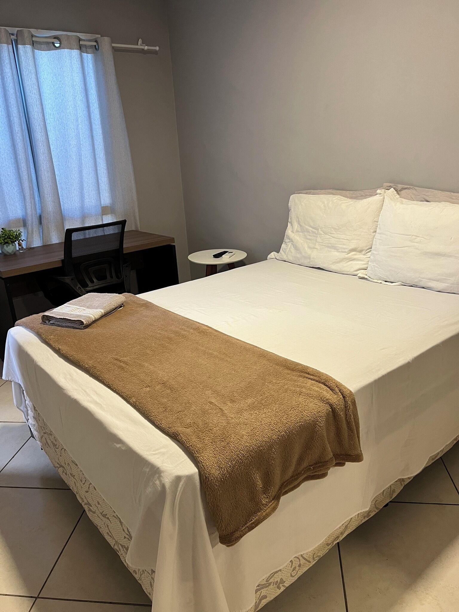 A neatly made bed with a brown blanket, a small desk with a chair, a round table, and a curtain.