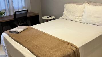 A neatly made bed with a brown blanket, a small desk with a chair, a round table, and a curtain.