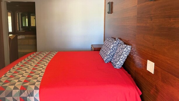 Habitación de hotel con una cama, cabecera de madera, colcha roja y almohada con motivos geométricos.