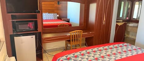 A compact hotel room with a bed, a desk, a chair, a small refrigerator, a microwave, and a mirror.
