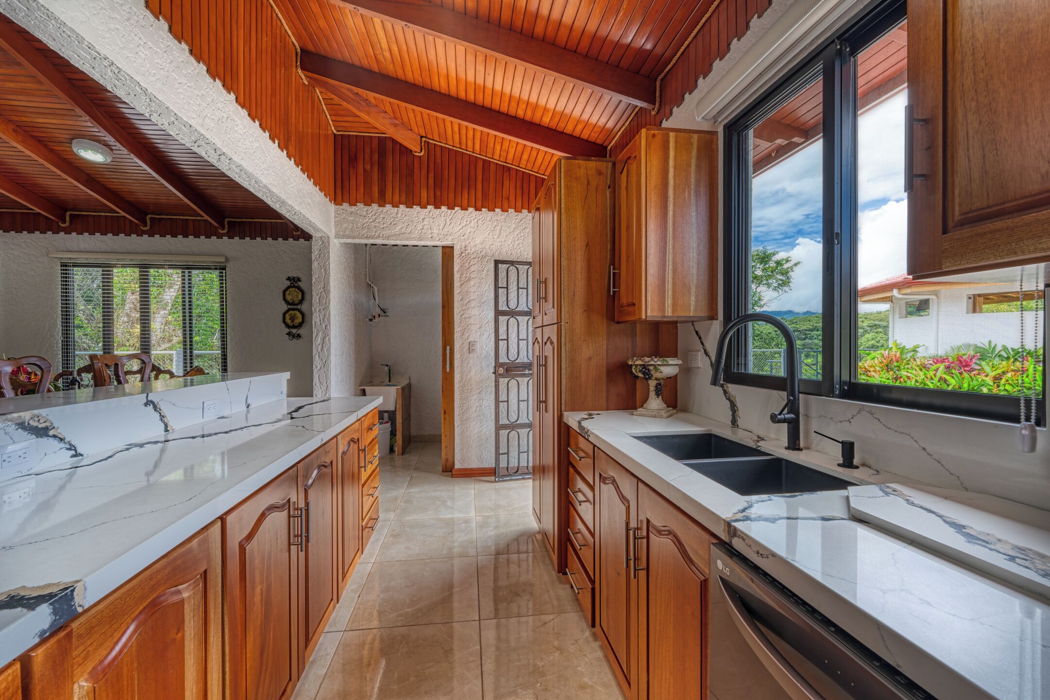 A kitchen with wooden cabinets, marble countertops, and a view of greenery outside.