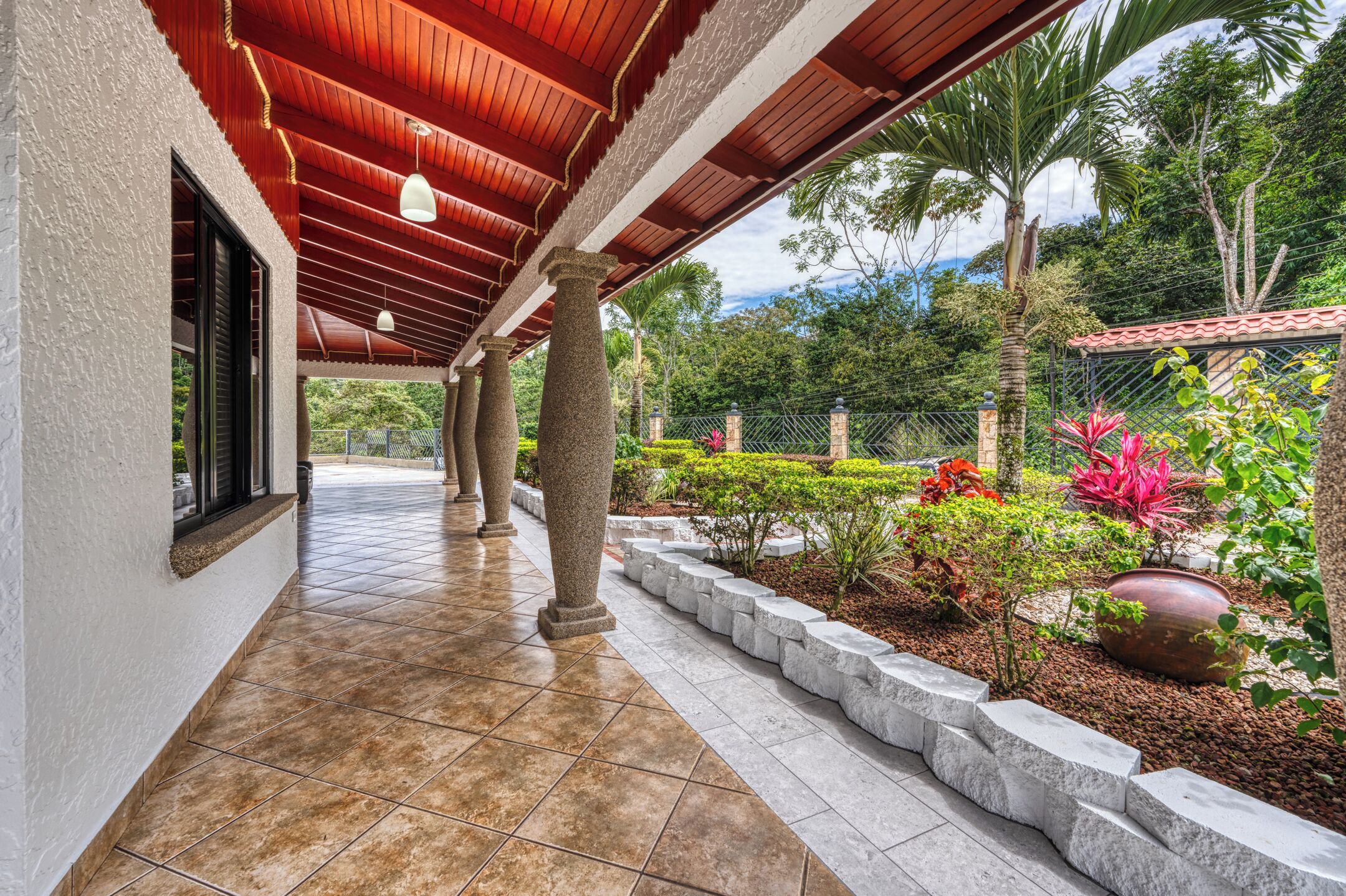 A covered outdoor corridor with stone columns, a tiled floor, and a garden with various plants and a large pot.