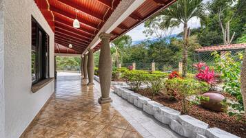 A covered outdoor corridor with stone columns, a tiled floor, and a garden with various plants and a large pot.