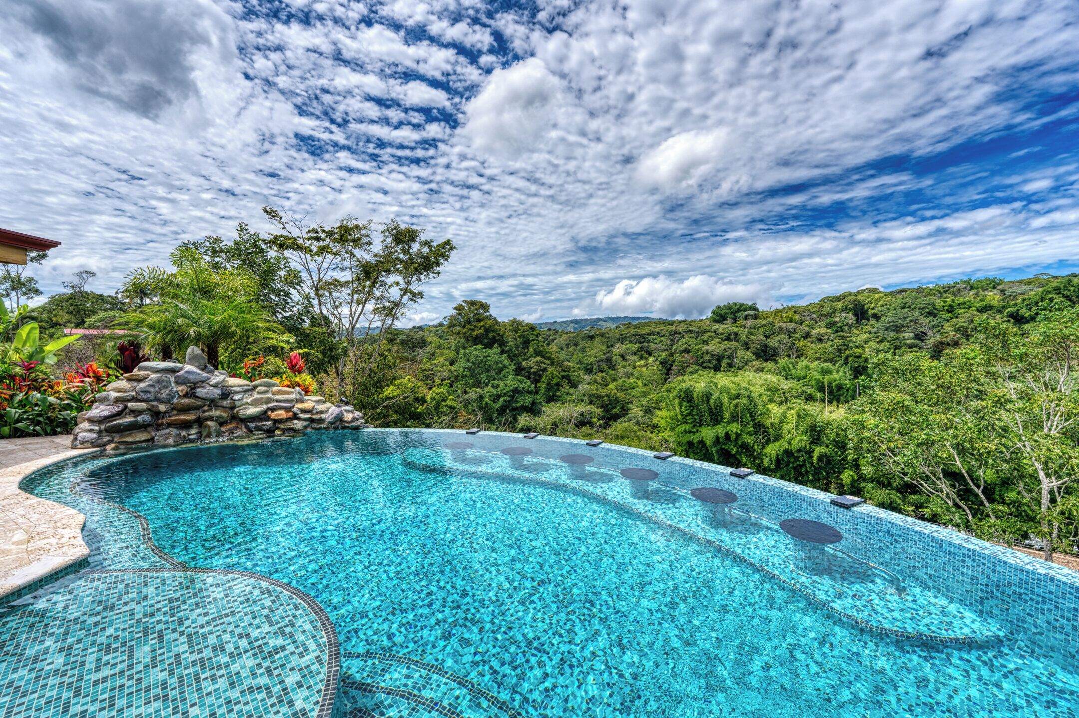 A clear blue swimming pool with a stone wall and tropical plants, surrounded by lush greenery and a cloudy sky.
