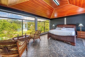 A bedroom with a large bed, two bedside tables, a chair, and a view of a lush landscape through large windows.