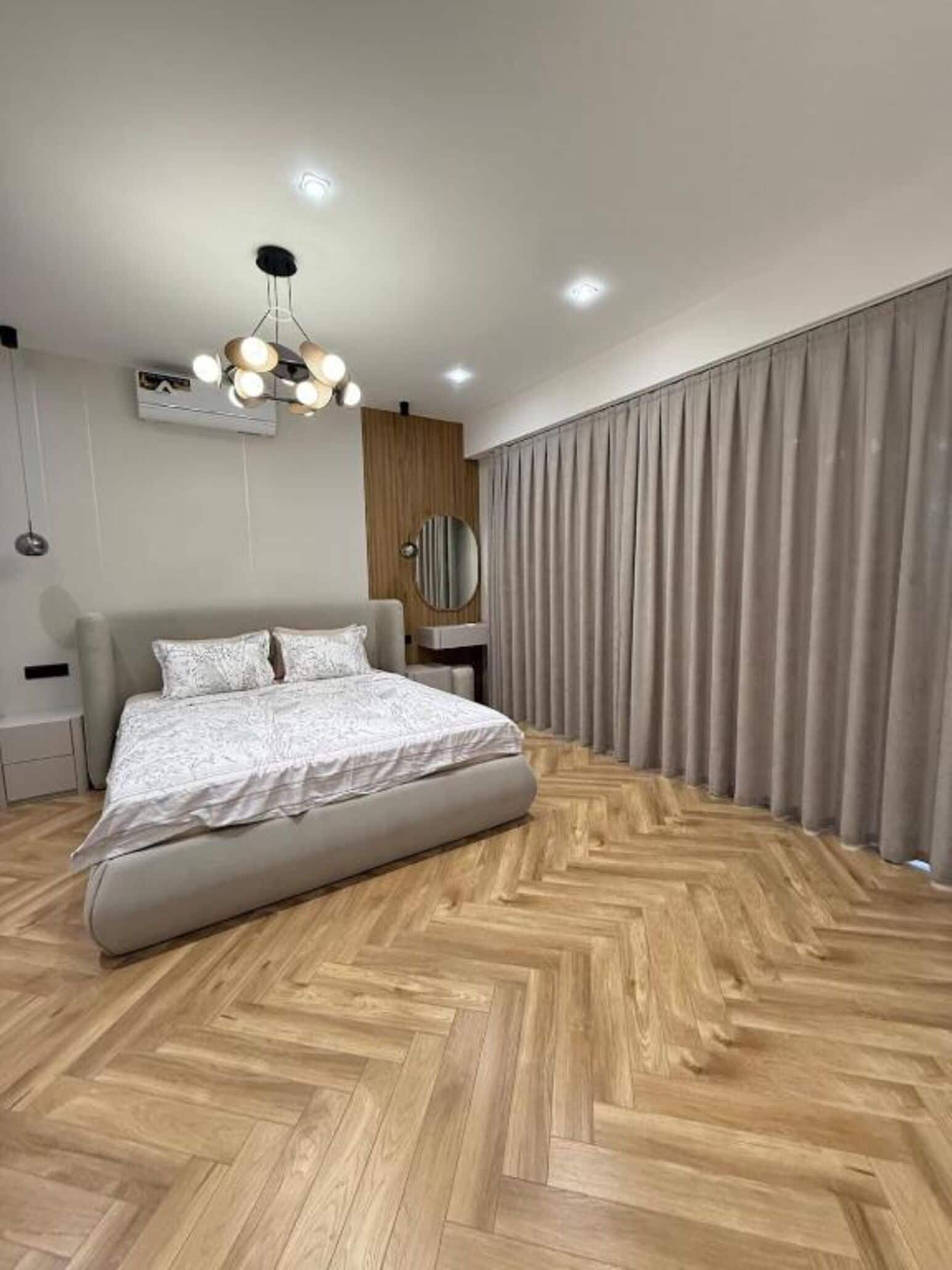 Une chambre à coucher moderne avec un grand lit, un parquet en bois disposé en chevrons, un luminaire au plafond et une fenêtre avec des rideaux.