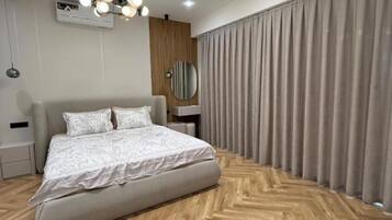 Une chambre à coucher moderne avec un grand lit, un parquet en bois disposé en chevrons, un luminaire au plafond et une fenêtre avec des rideaux.